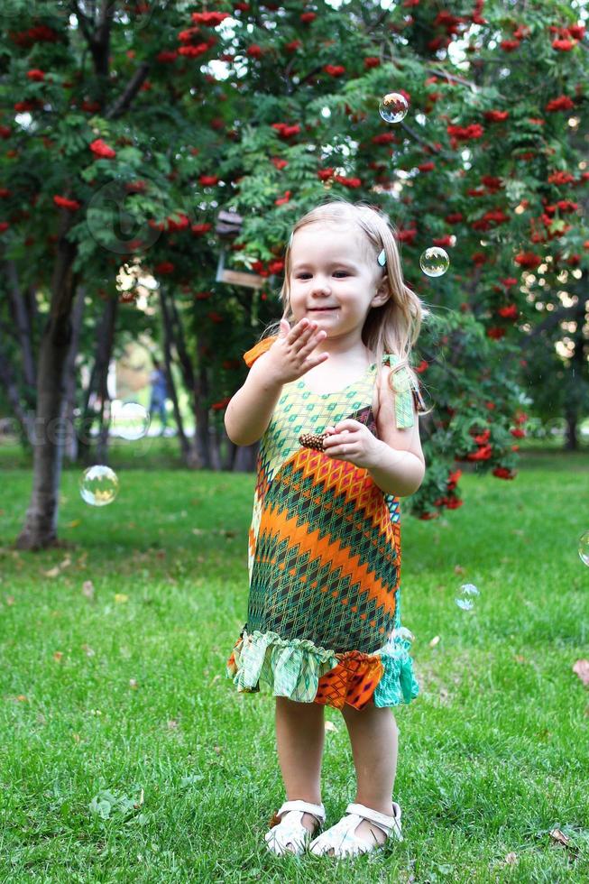 una linda niña sonriente con coloridos vestidos de verano está jugando con pompas de jabón en el parque de verano. foto