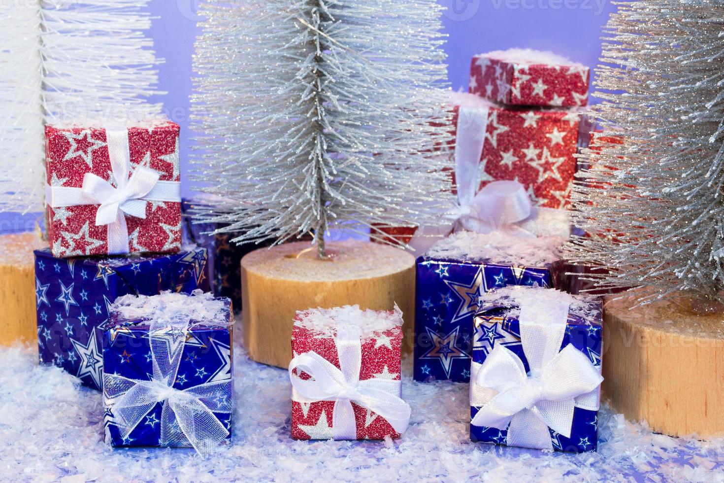 Gift boxes wrapped in shiny blue and red paper with artificial snow and Christmas trees on purple background. photo