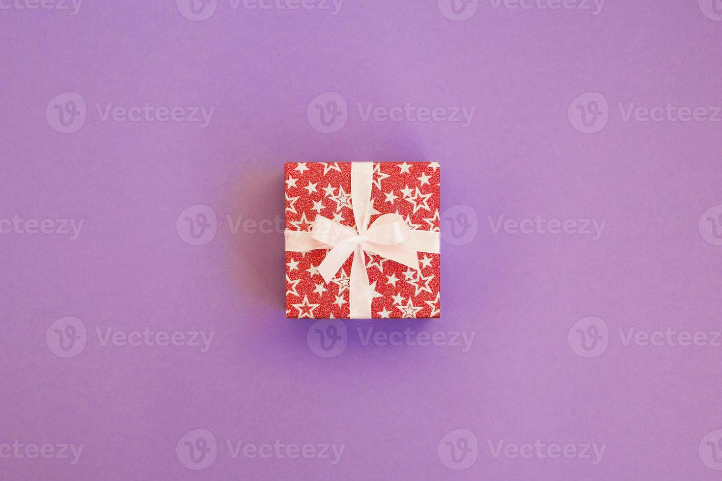 Gift box wrapped in red shiny paper with white ribbon on purple background. Mockup with copy space. photo
