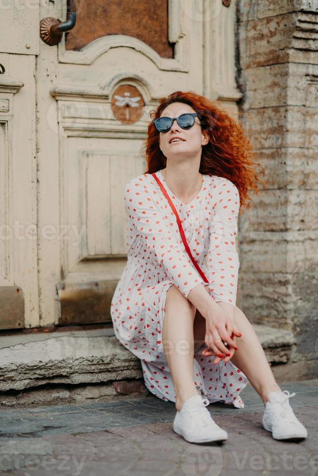 Pleased red haired female with curly hair, wears sunglasses and dress, poses near door of ancient building, holds hands together, enjoys rest in open air, likes viewing old historic famous places photo
