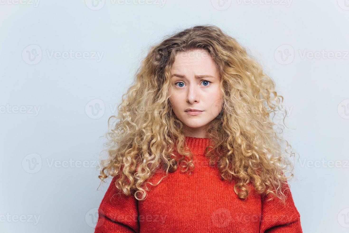 Photo of displeased unhappy young woman with curly bushy blonde hair, frowns face in bewilderment, wears casual clothing, models against white studio background. People and disappointment concept