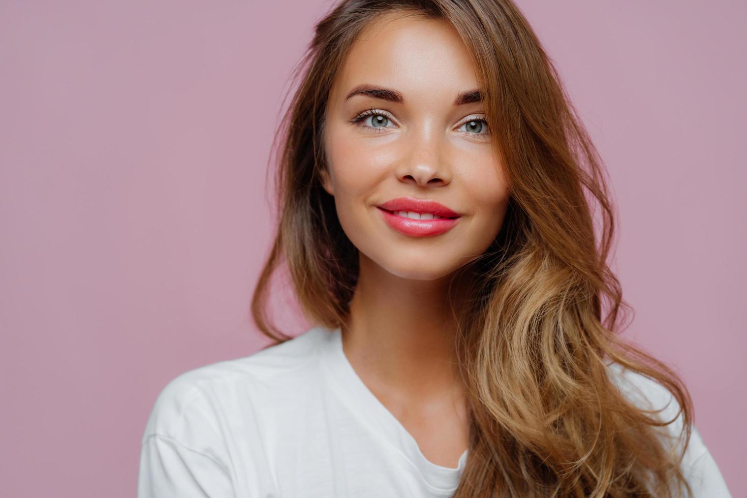 Horizontal cropped image of pleasant looking female model has tender smile, wears minimal makeup, has long wavy hair, looks at camera with satisfaction, models over violet wall, copy space for promo photo