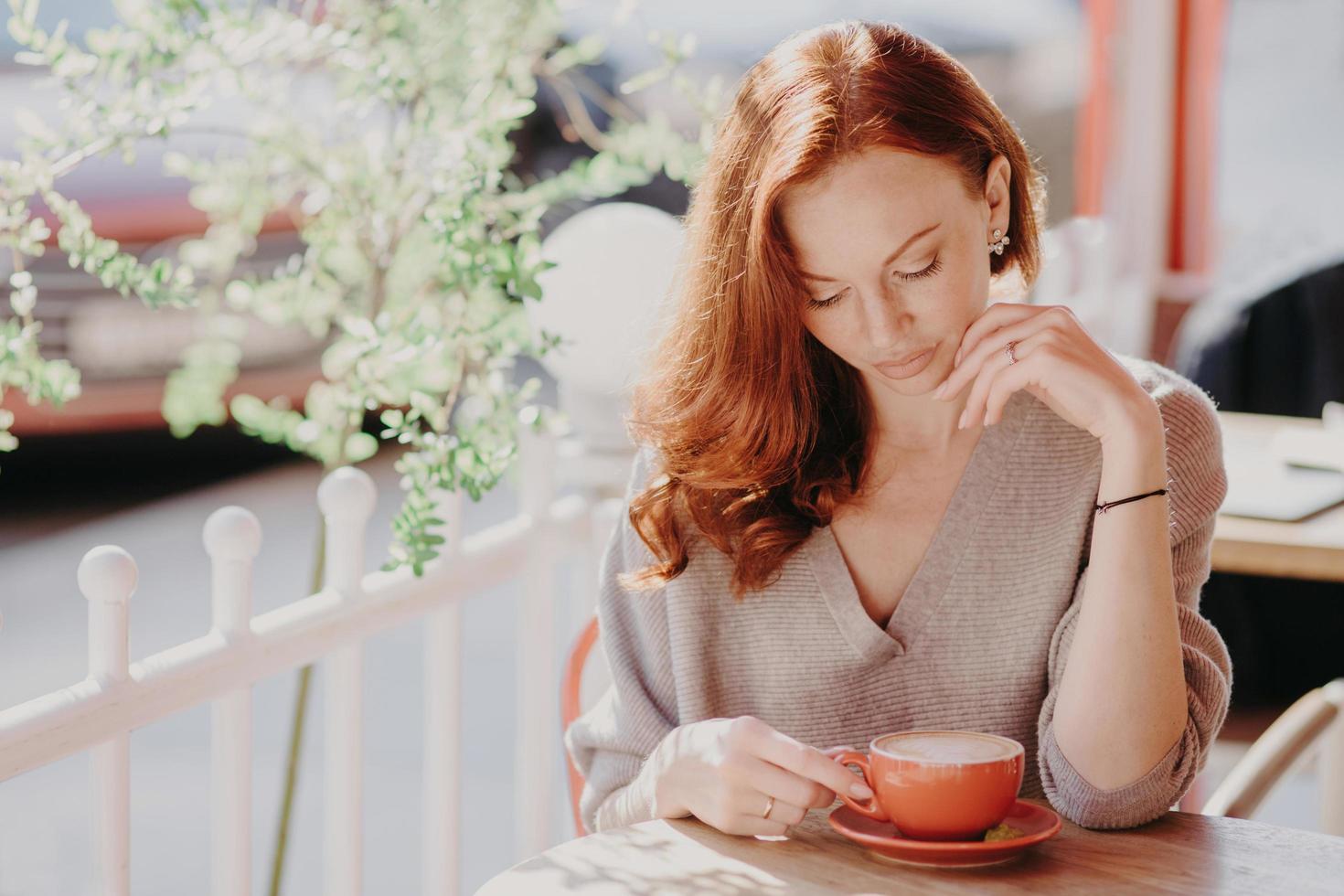 Charming red haired European woman drinks cappuccino or coffee on terrace cafe, has calm facial expression, wears brown sweater, has make up, enjoys good drink. People and lifestyle concept. photo