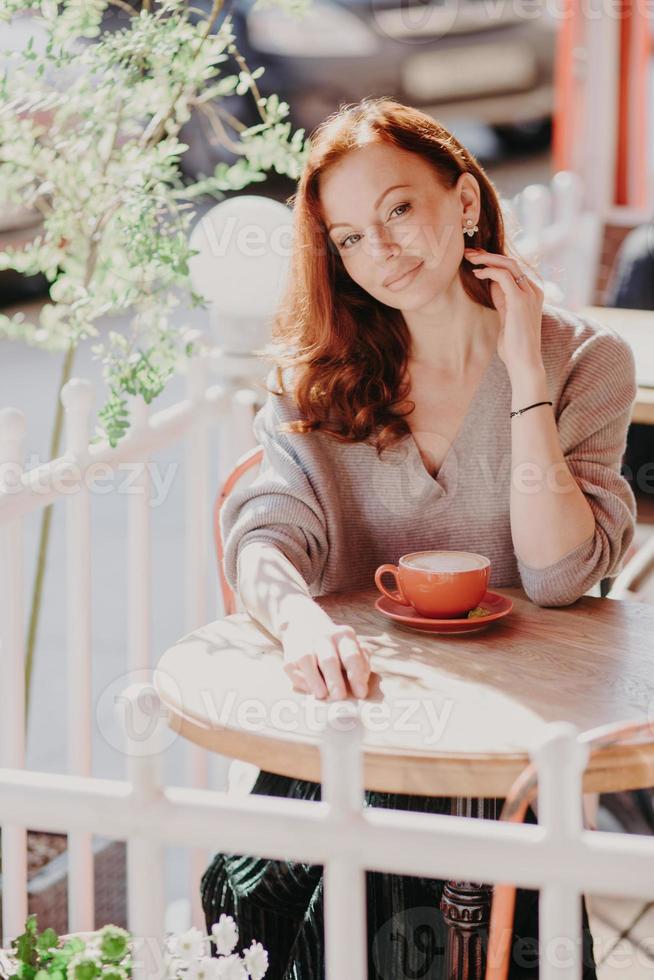 Photo of good looking ginger young woman in spectacles watches tutorial video online, poses in comfortable couch in modern apartment, dressed in elegant outfit, enjoys free internet connection