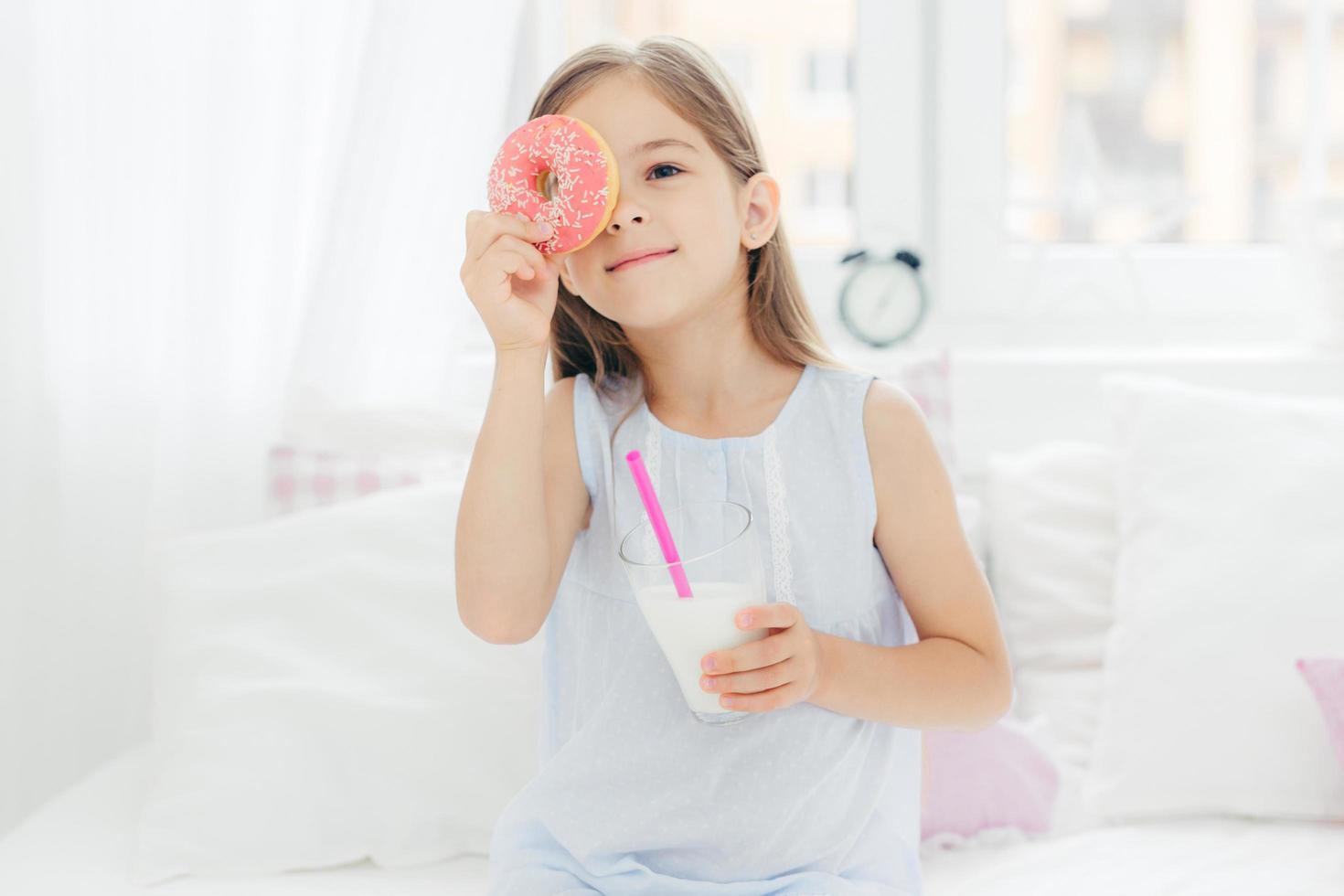 Cheerful little female child poses in bedroom wih delicious doughnut and milk shake, sits on comfortable bed, has fun in morning, going to have tasty breakfast. Children and bed time concept photo