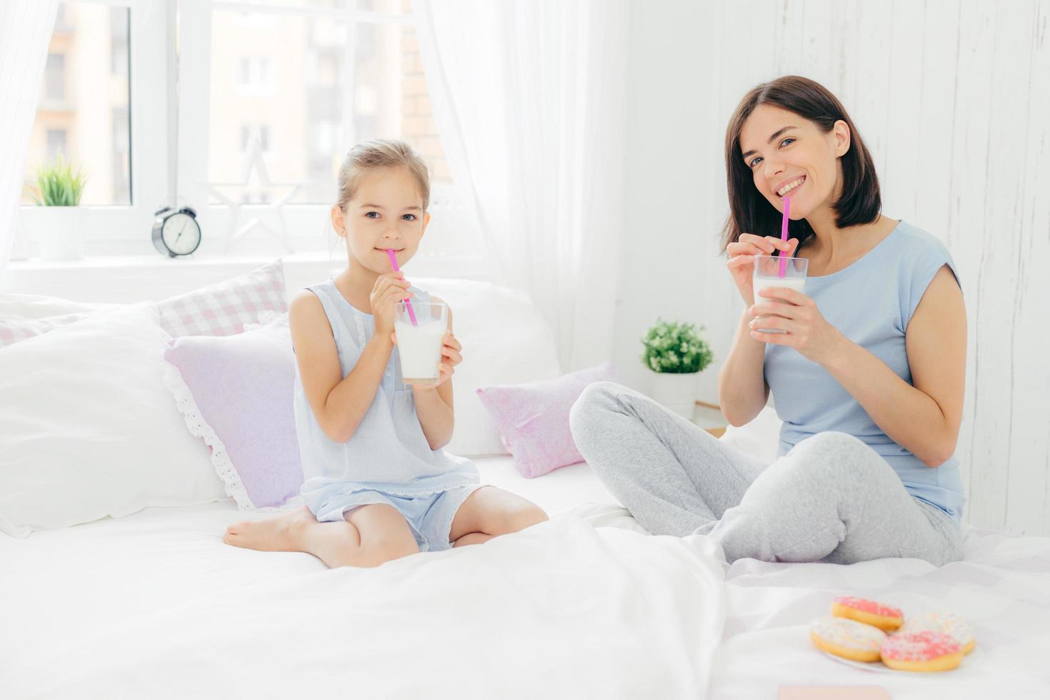 Positive mother and daughter start their day with delicious breakfast, drink cocktail with tasty sweet doughnuts, sits on white bed, have happy expression. People, morning and awakening concept photo