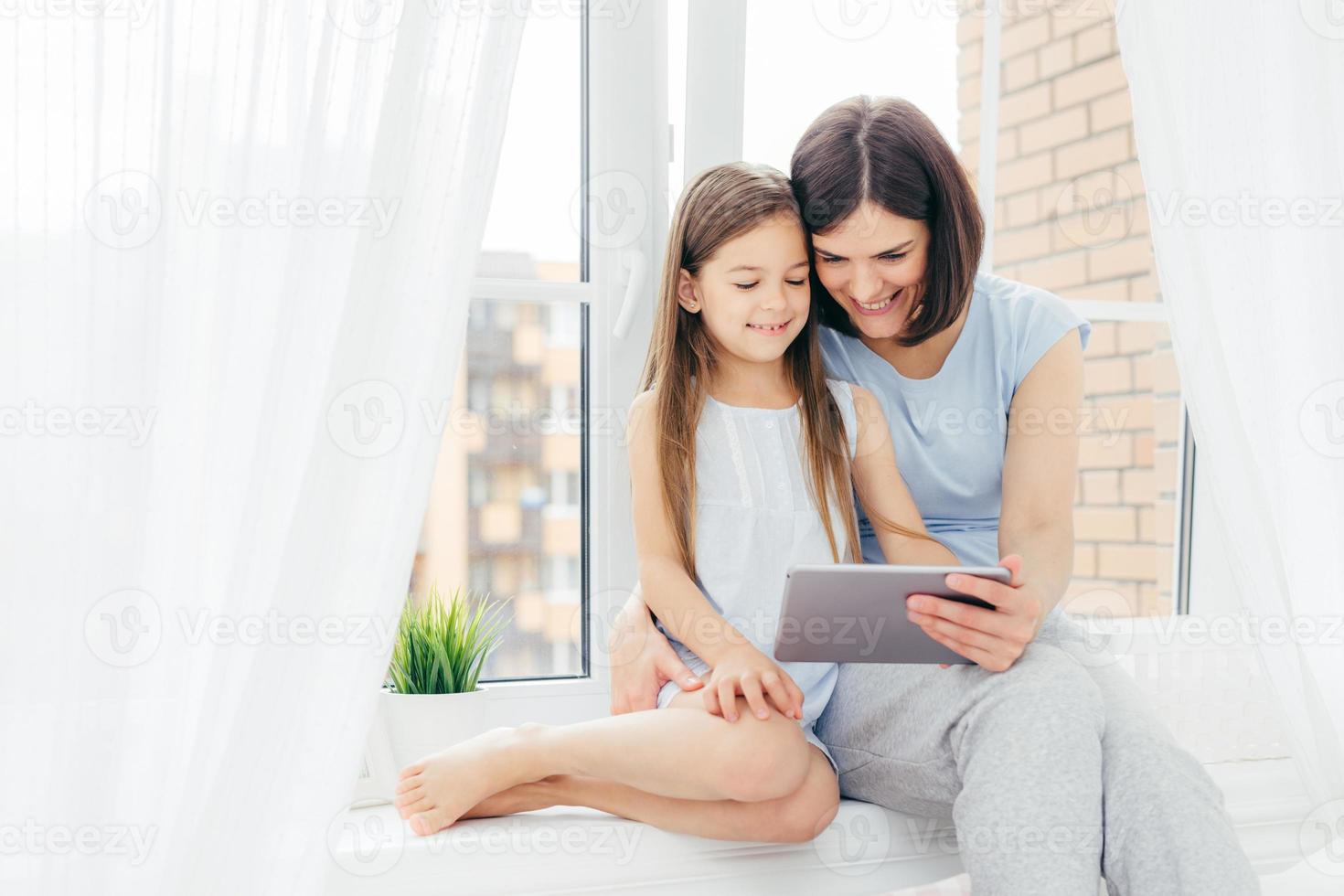 People, technology, family, children concept. Positive young other and her small daughter sit on window sill, hold digital tablet, watch interesting cartoon, pose near window with white curtains photo