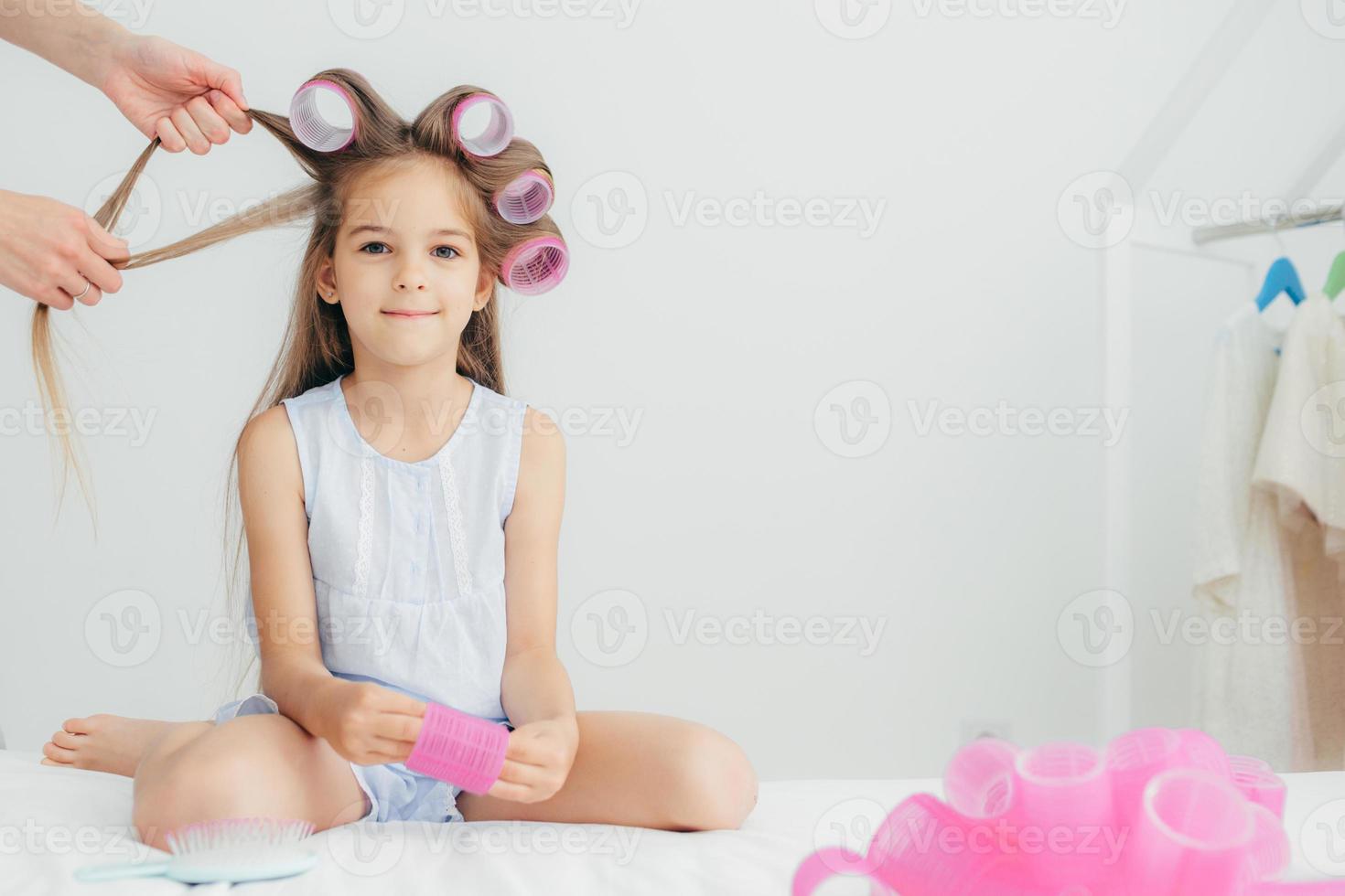 Attractive female child has curlers on head, going to have wonderful hairstyle, recieves beauty lessons from affectionate mother, isolated over white background. Children and beauty concept. photo