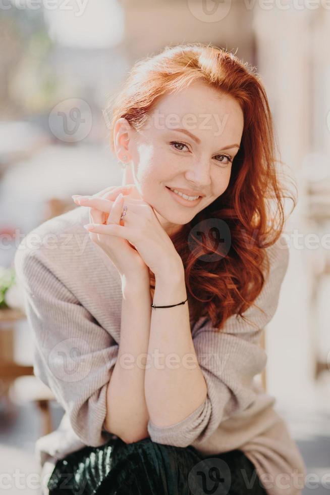Image of lovely redhead female model smiles happily, has healthy skin, make up, keeps hands together, dressed in oversized jumper, poses outside, smiles gladfully. Having nice day or weekend photo