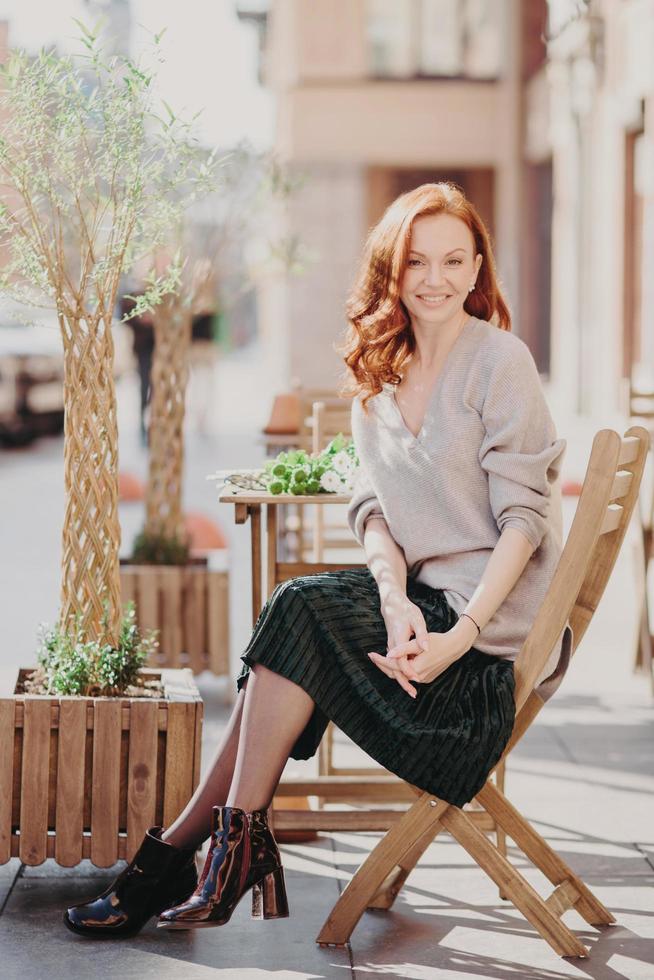 Photo of beautiful red haired woman dressed in stylish clothes, sits in open air, poses at cafeteria, has pleasant appearance, enjoys leisure time, smiles positively. People and lifestyle concept