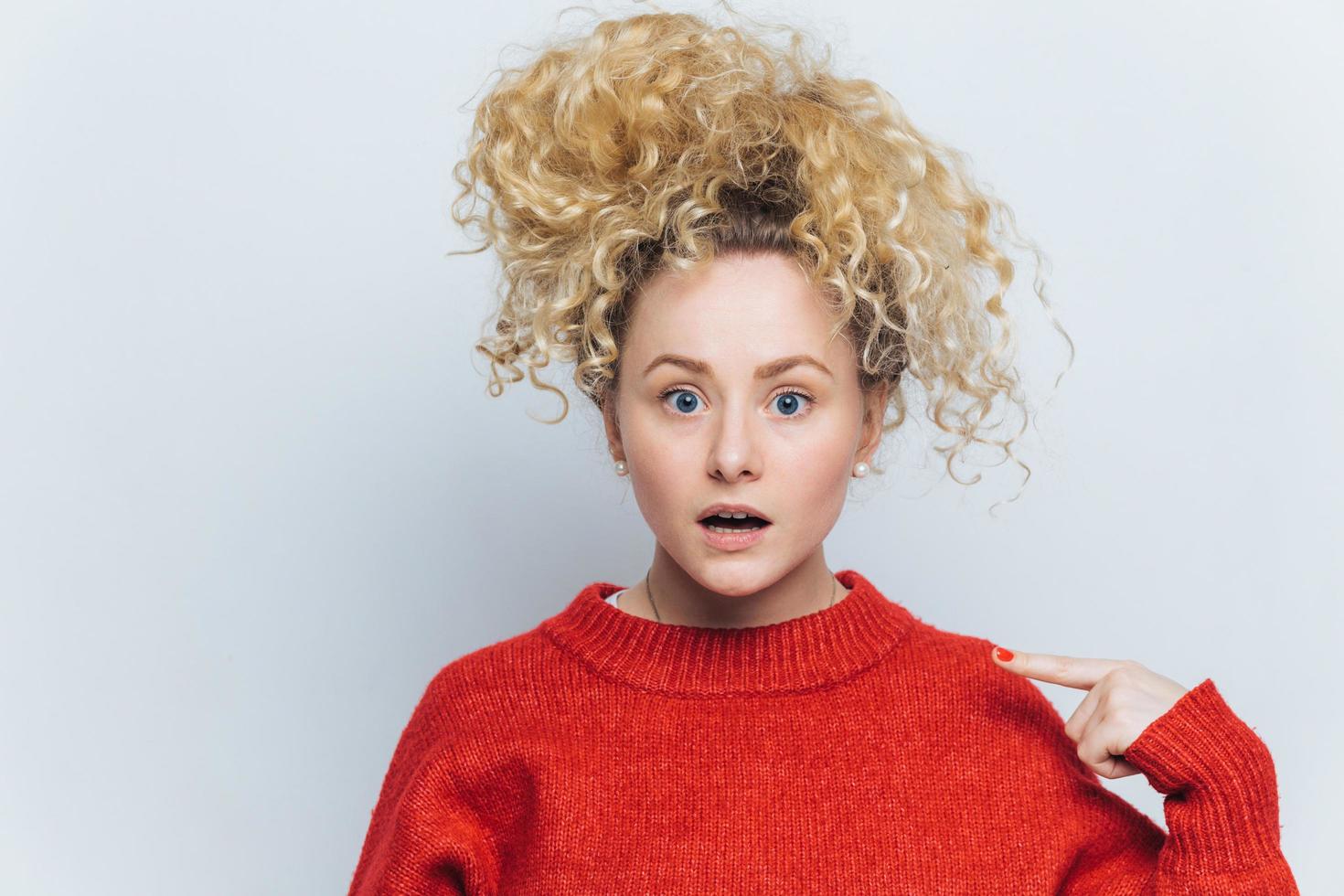 Shocked emotional amazed female stares at camera, indicates at sweater for your advertisment or information, isolated over white background. Surprised curly young woman isolated on white wall photo