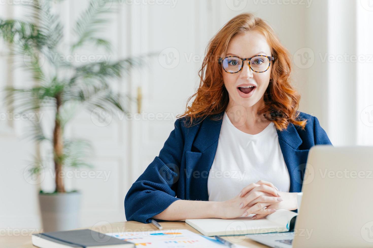 una empleada pelirroja sorprendida tiene trabajo, desarrolla una nueva estrategia de negocios, posa frente a una computadora portátil abierta, posa en el espacio de trabajo contra el interior de la oficina. mujer desarrolladora de software en el trabajo. foto