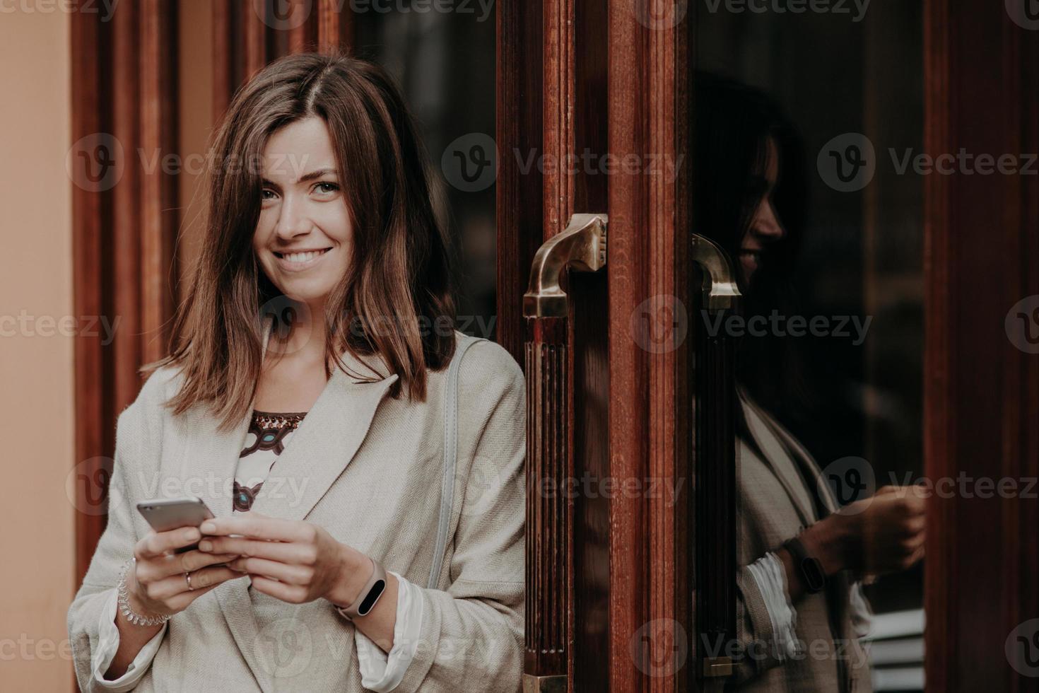 Cheerful glad female with dark straight hair, uses smart phone, dressed in elegant clothing, looks positively at camera, enjoys online communication, checks route, sends messages, reads notification photo