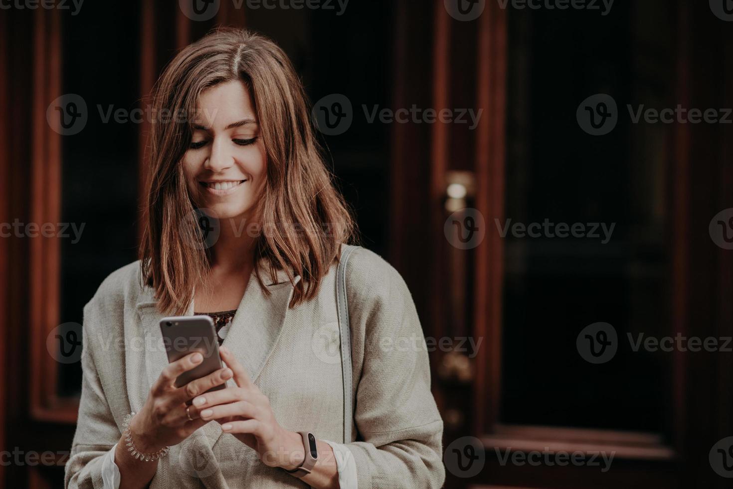 Horizontal shot of pretty cute brunette female model shares multimedia files via cell phone, dressed in elegant clothes, reads positive news in internet website, connected to wireless internet. photo