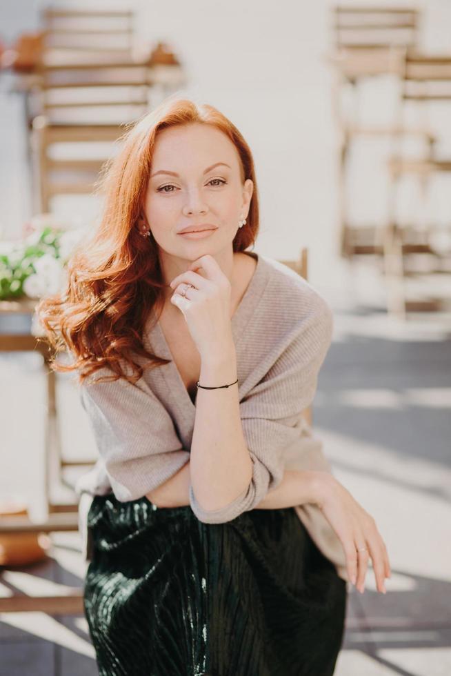 Image of lovely woman with ginger hair, has make up, keeps hand under chin, looks directly at camera, wears stylish clothes, poses over street background, has confident expression. Beauty and rest photo