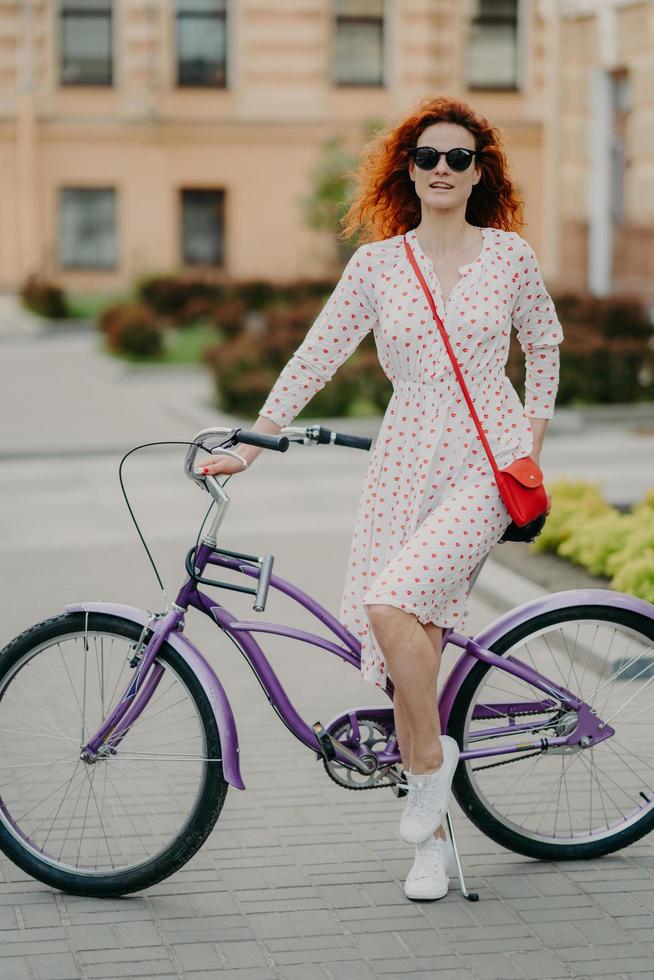 Vertical shot of beautiful red haired female model has rest after covering long distance on bicycle, wears sunglasses, summer dress and white sneakers, enjoys sport and exploring city streets photo