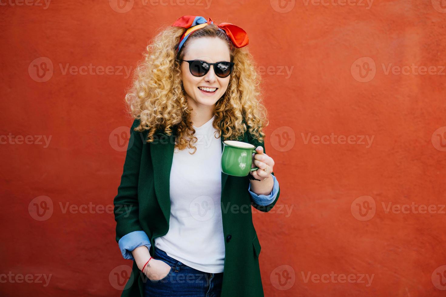 atractiva joven europea con diadema con elegante chaqueta y gafas sosteniendo una taza de capuchino con una expresión feliz mientras se enfrenta a un fondo naranja. gente, concepto de estilo de vida foto