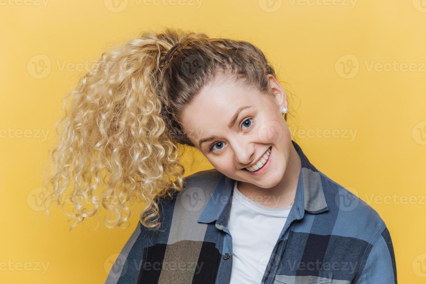 Funny cute female model with curly blonde pony tail, has positive smile, has fun alone indoor against yellow background, wears casual checkered shirt, being in good mood. Facial expressions. photo