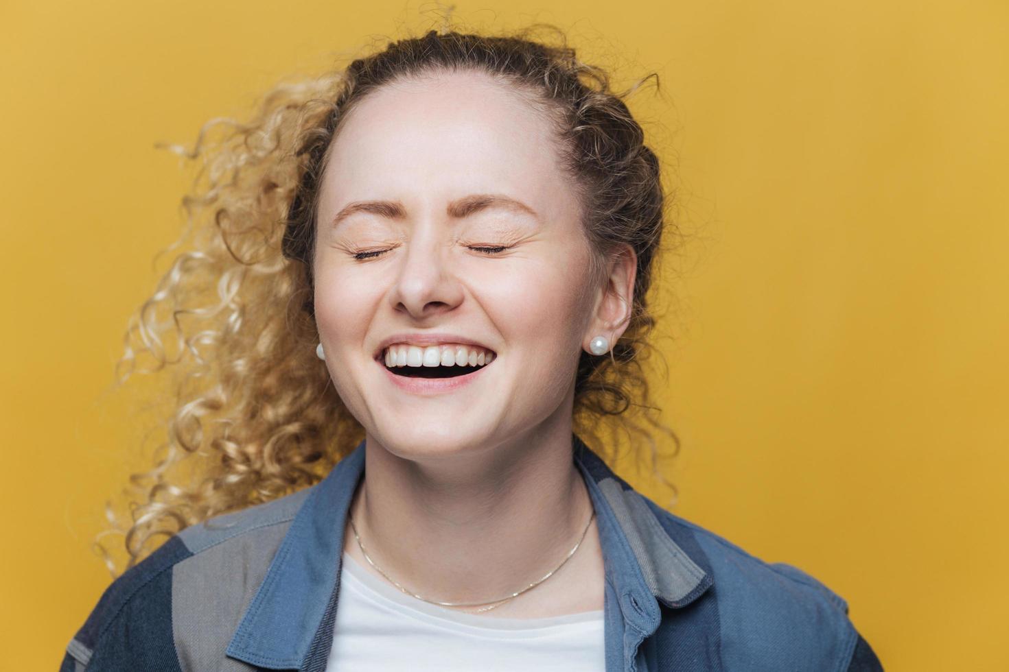 Overjoyed beautiful female laughs joyfully as hears good joke, smiles broadly, keeps eyes shut, has curly hair, isolated over yellow background. Cute young woman poses indoor, expresses happiness photo