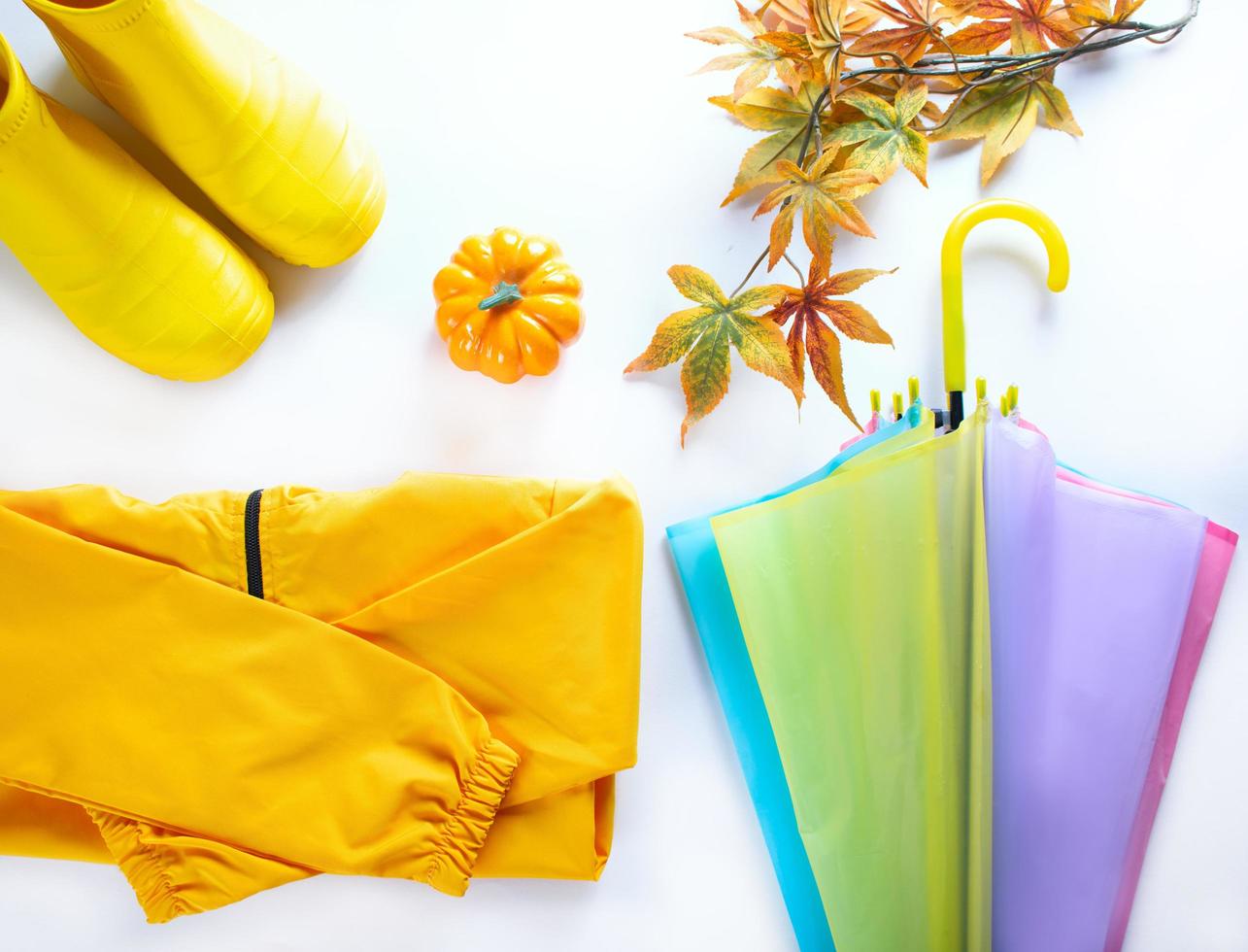 Colorful umbrella, yellow rubber boots, raincoat and autumn leaves on light background. Outfit for kids. Top view, flat lay photo