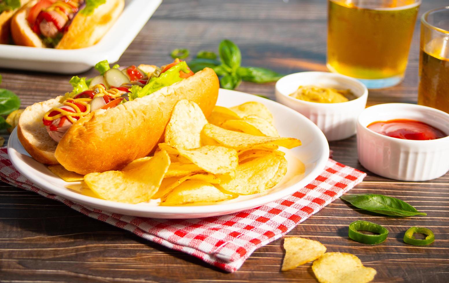 Delicious homemade hotdogs with dips sauces, chips and glasses of beer on wooden table. Top view. Copy space. photo