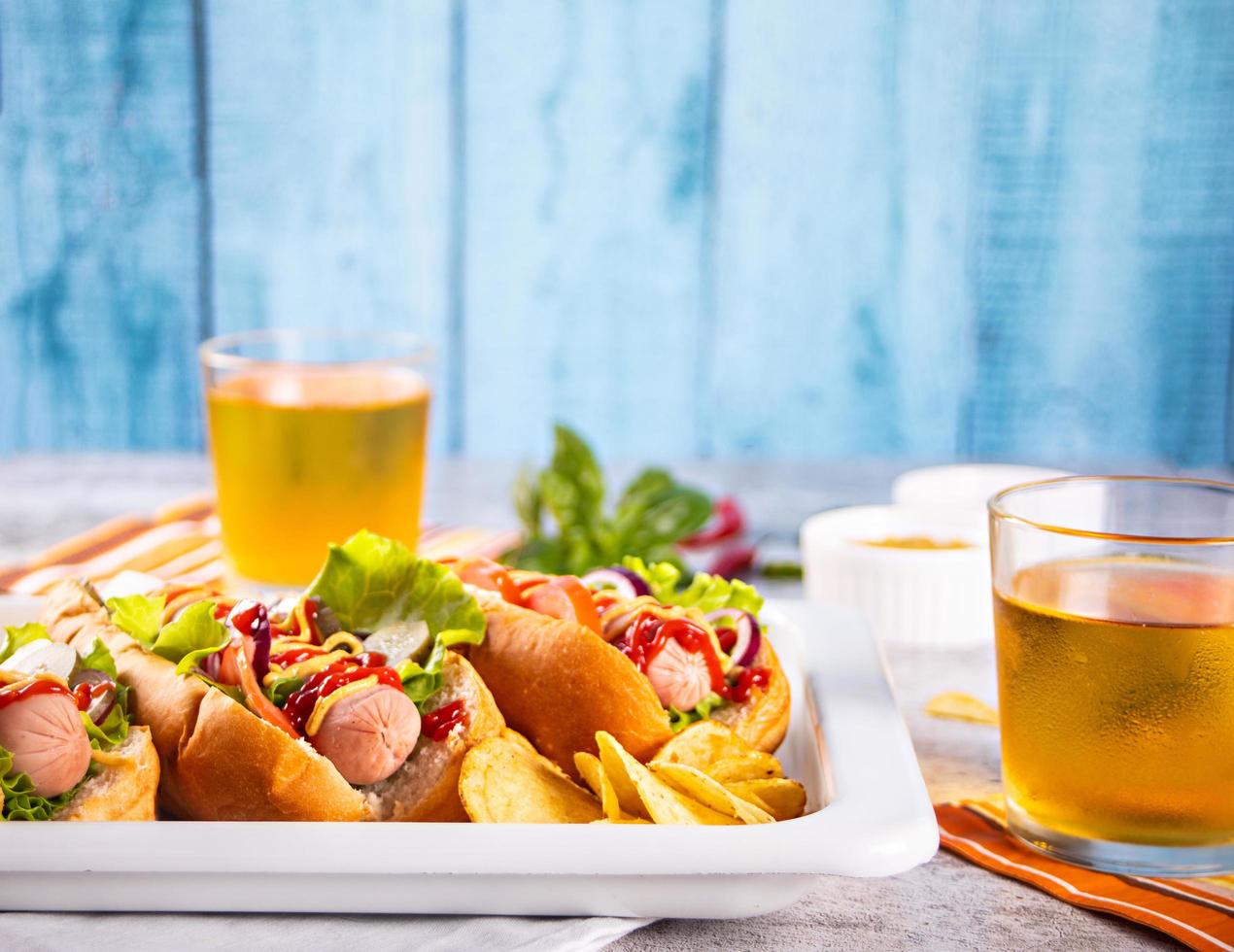 Delicious homemade hotdogs with dips sauces, chips and glasses of beer on wooden table. Top view. Copy space. photo