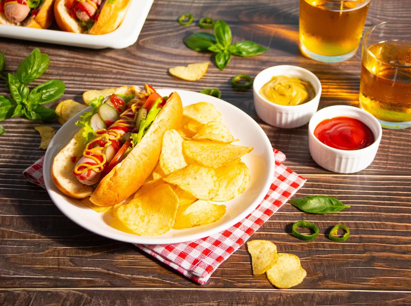 deliciosos perritos calientes caseros con salsas, patatas fritas y vasos de cerveza sobre una mesa de madera. vista superior. copie el espacio foto