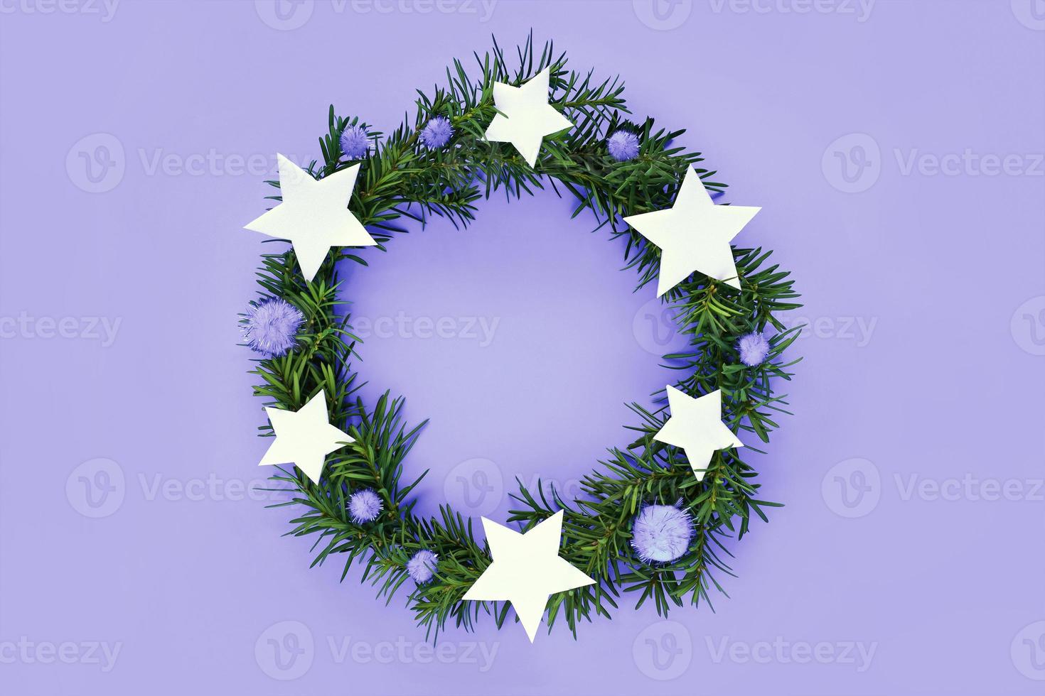 Christmas wreath made of fir-tree branches on very peri trend color background. Wreath decorated violet fluffy pom poms and white stars. Flat lay, top view, copy space. Color of new year 2022 concept. photo