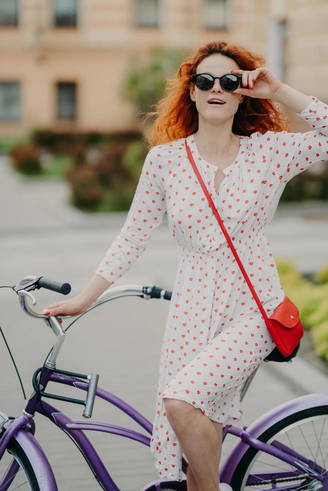 Street style and active rest concept. Curly red haired woman keeps hand on frame of sunglasses, wears white dress, focused into distance, waits for friend, going to have outdoor trip on bicycle photo
