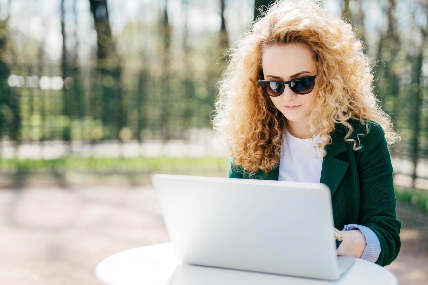 una estudiante seria que tiene el pelo rubio esponjoso con una chaqueta verde y gafas de sol sentada al aire libre haciendo su trabajo de diploma disfrutando de un hermoso clima soleado. moda, belleza, concepto juvenil. foto