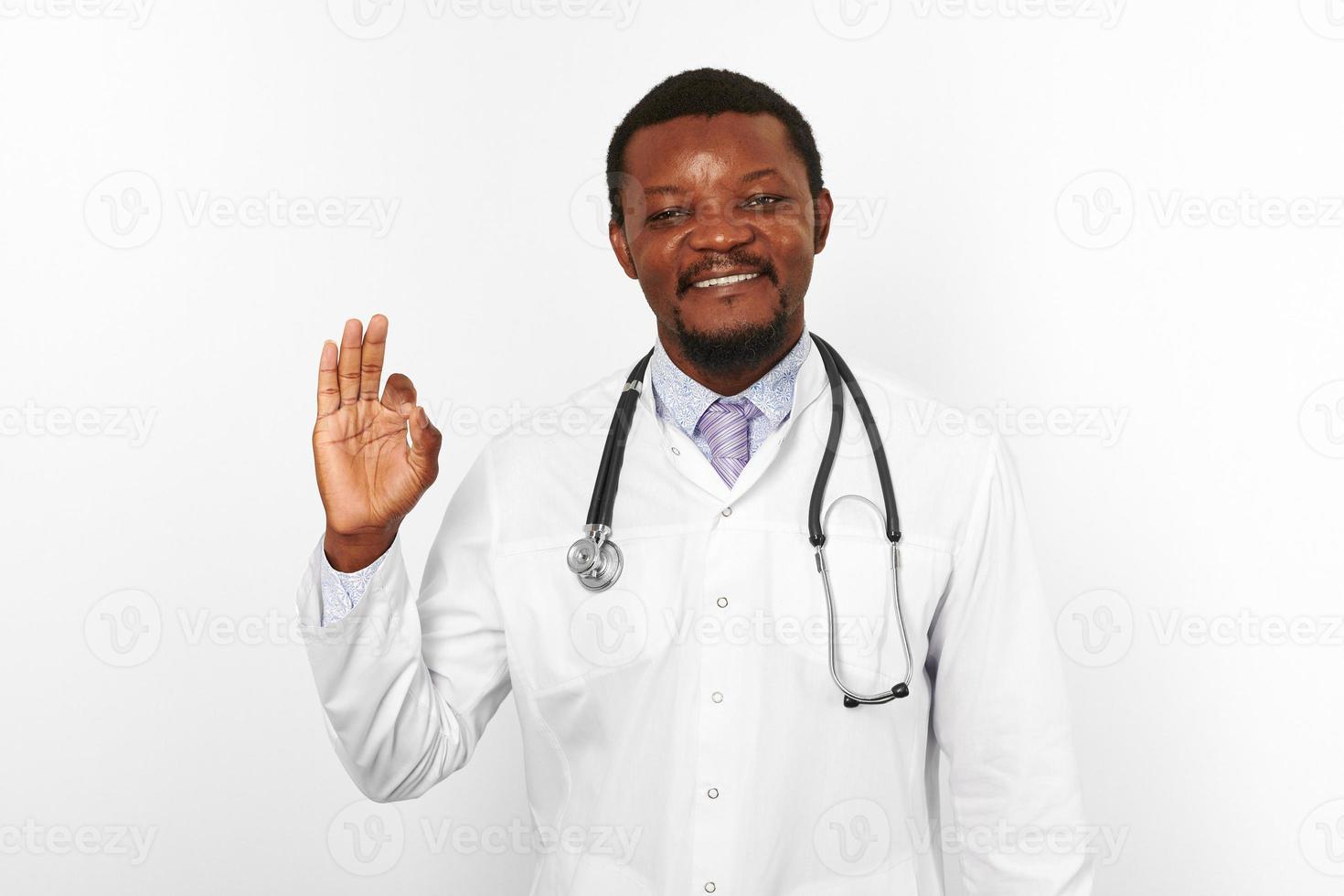 Smiling black bearded doctor man in white robe with stethoscope shows OK gesture, white background photo