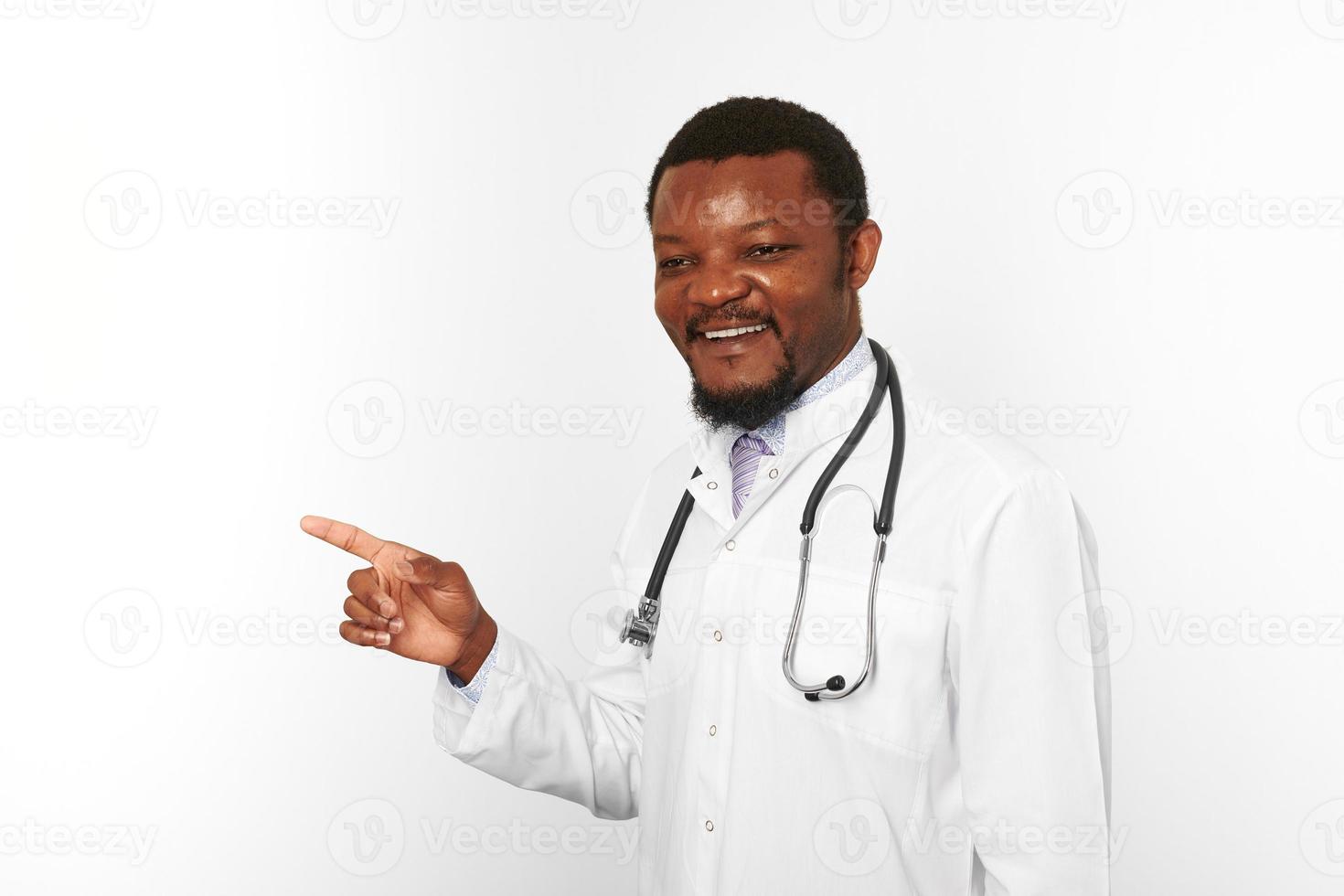 Smiling black bearded doctor man in white coat with stethoscope points finger, white background photo