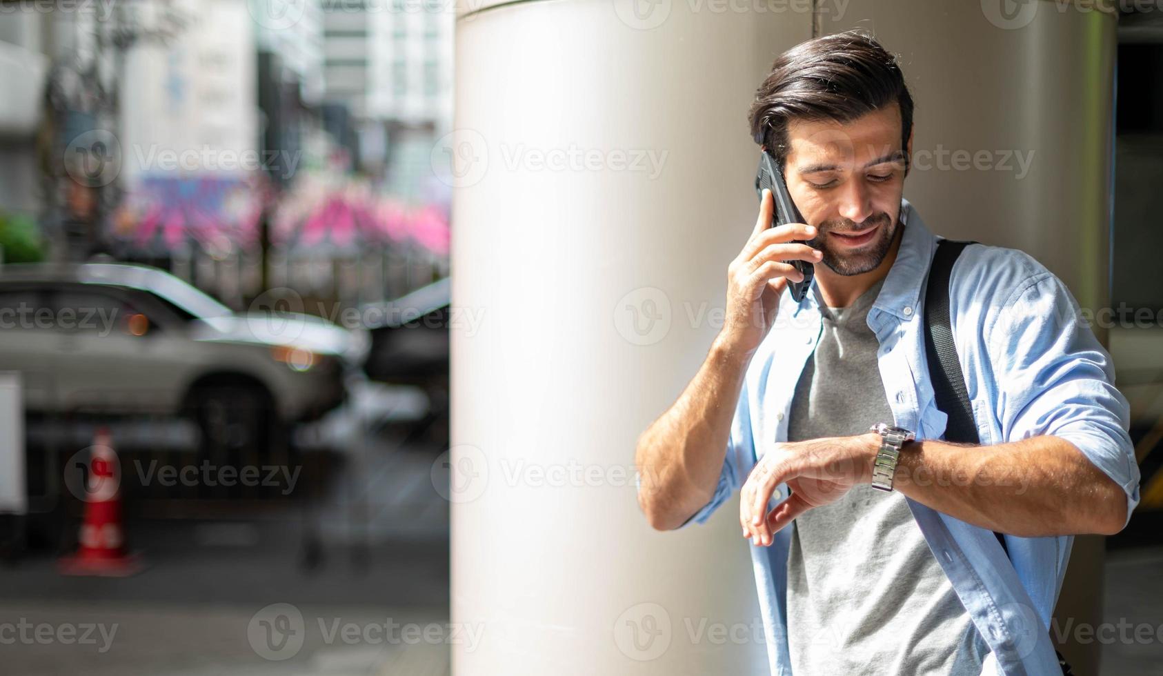 joven caucásico mirando la hora y llamando por teléfono celular mientras espera algo frente a la oficina. foto