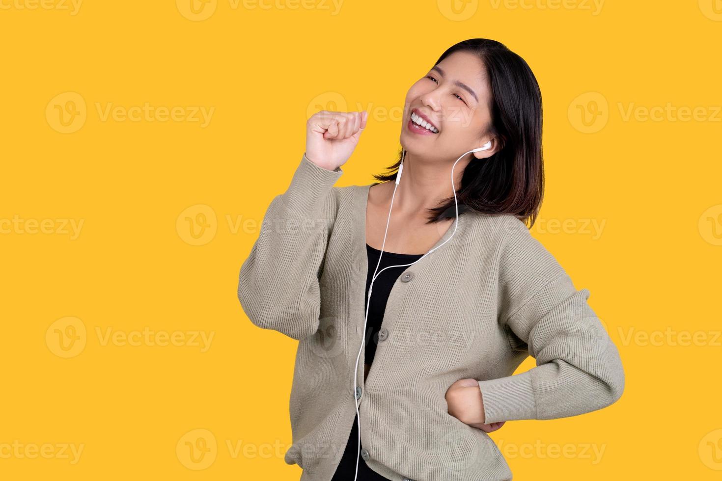 mujer hermosa asiática feliz cantando su canción favorita. lleva auriculares blancos aislados en un fondo amarillo brillante. foto