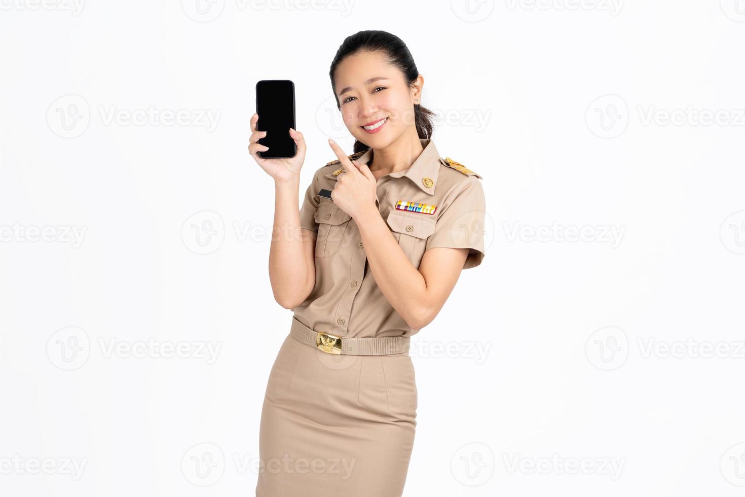 Smiling young Asian female teacher demonstrates promo on phone isolated on white background. She is pointing finger at smartphone and smiling. photo
