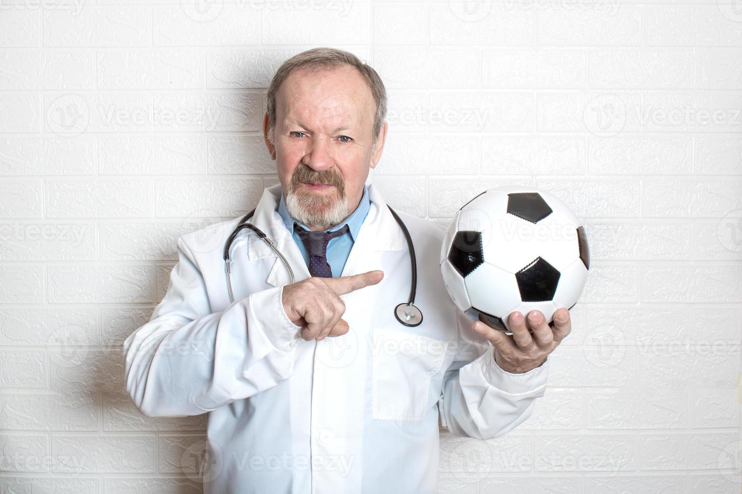 un médico anciano serio sostiene una pelota de fútbol en una mano y la señala con la otra. Esta es una fuerte recomendación para practicar deportes a cualquier edad. foto