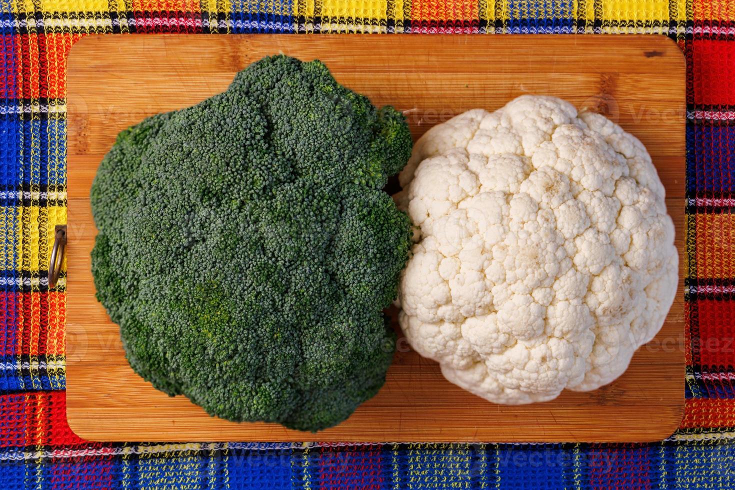 a head of broccoli and a head of cauliflower lie on a cutting board on the table with colorful towel underneath photo