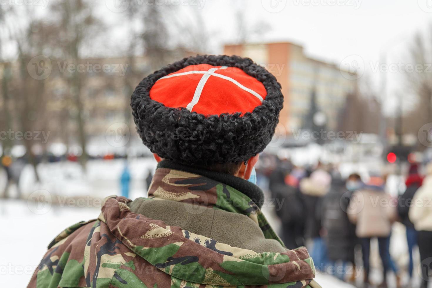 man in camouflage jacket and cossack hat with white cross on red watching blurry crowd of people photo