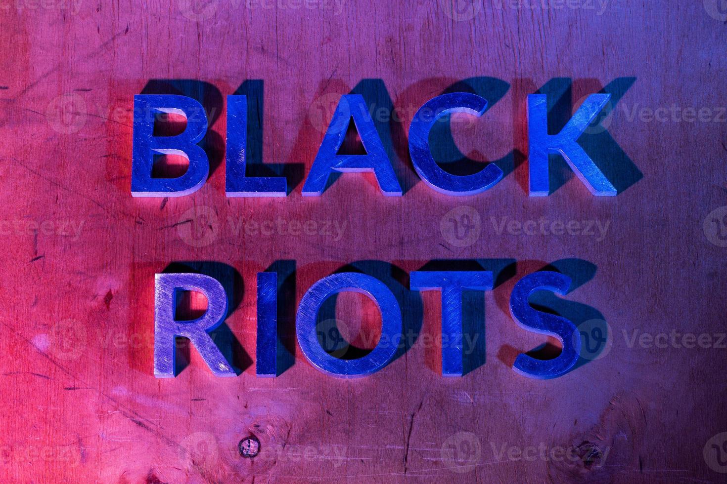 The words black riots laid with metal letters on flat plywood surface under red and blue police lights. Concept. photo