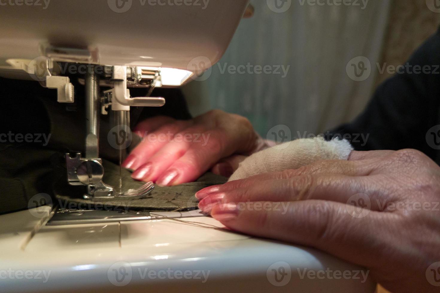 a hand of aged womans with a bandaged finger sews with a sewing machine photo