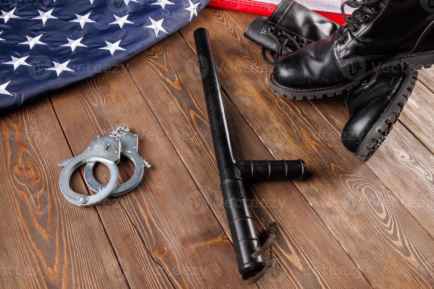 esposas de metal plateado, botines negros y bastón de policía cerca de la bandera estadounidense en la superficie de madera foto