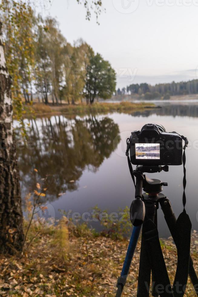 black digital camera on tripod shooting early foggy morning landscape at autumn lake with selective focus photo