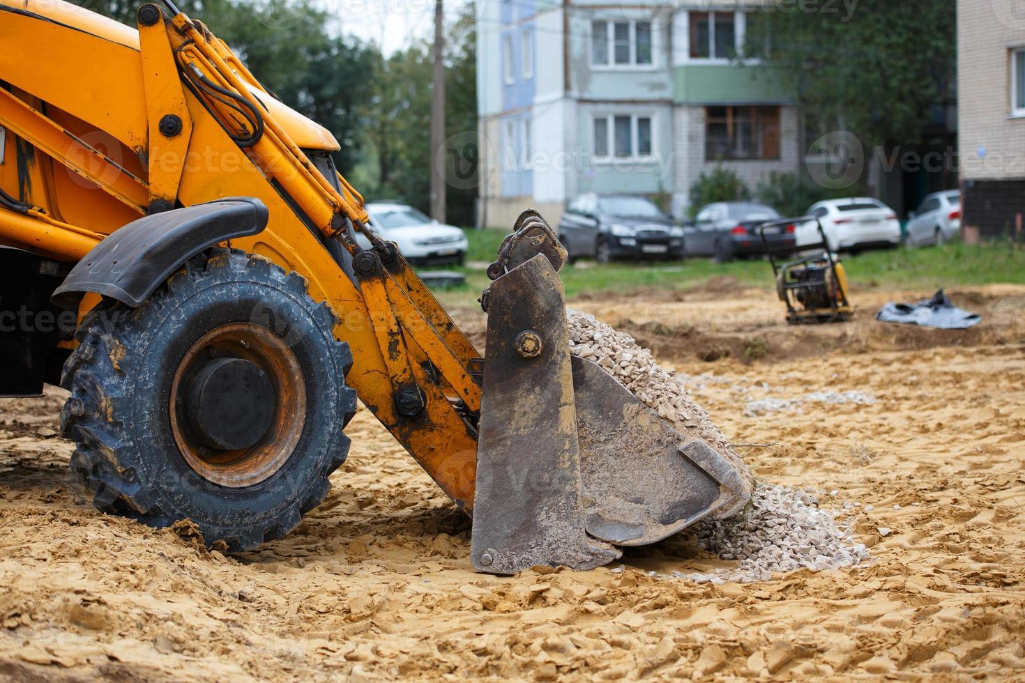 Cubo de excavadora de ruedas lleno de grava frente a casas y automóviles borrosos foto