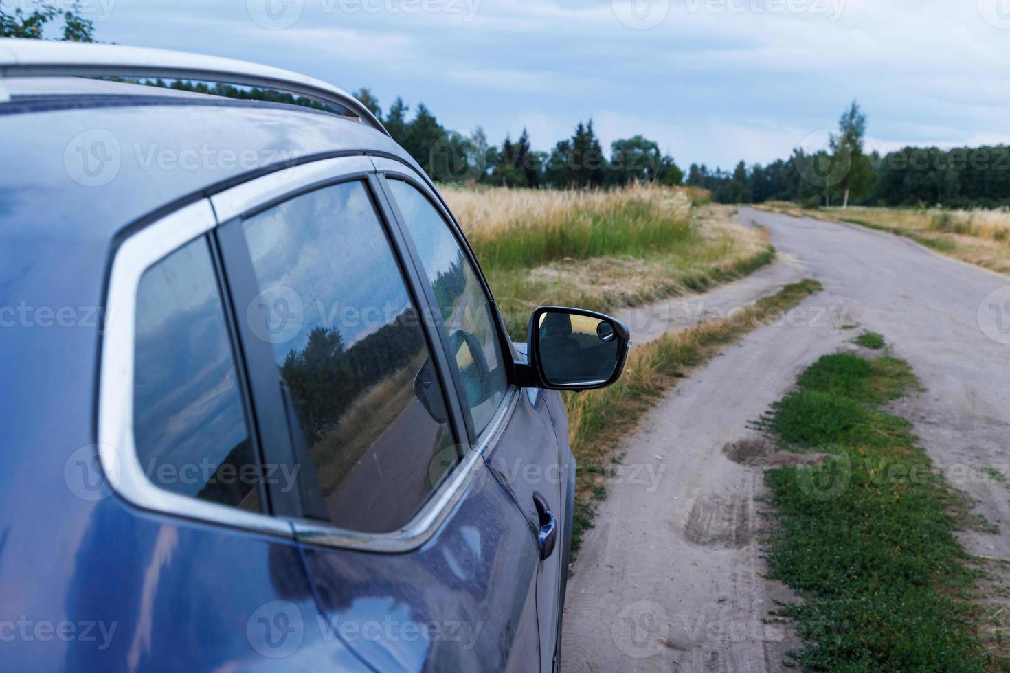 violet car on dry dirt road at summer evening with dry tall yellow grass and dark green forest in blurry background photo