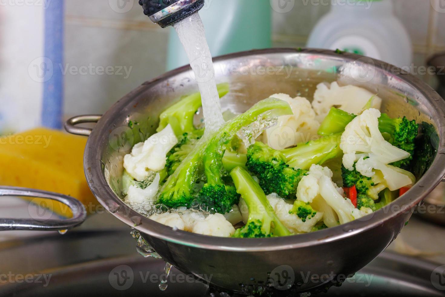 tap water flows over chopped cauliflower and broccoli in a colander above the sink while rinsing vegetables photo