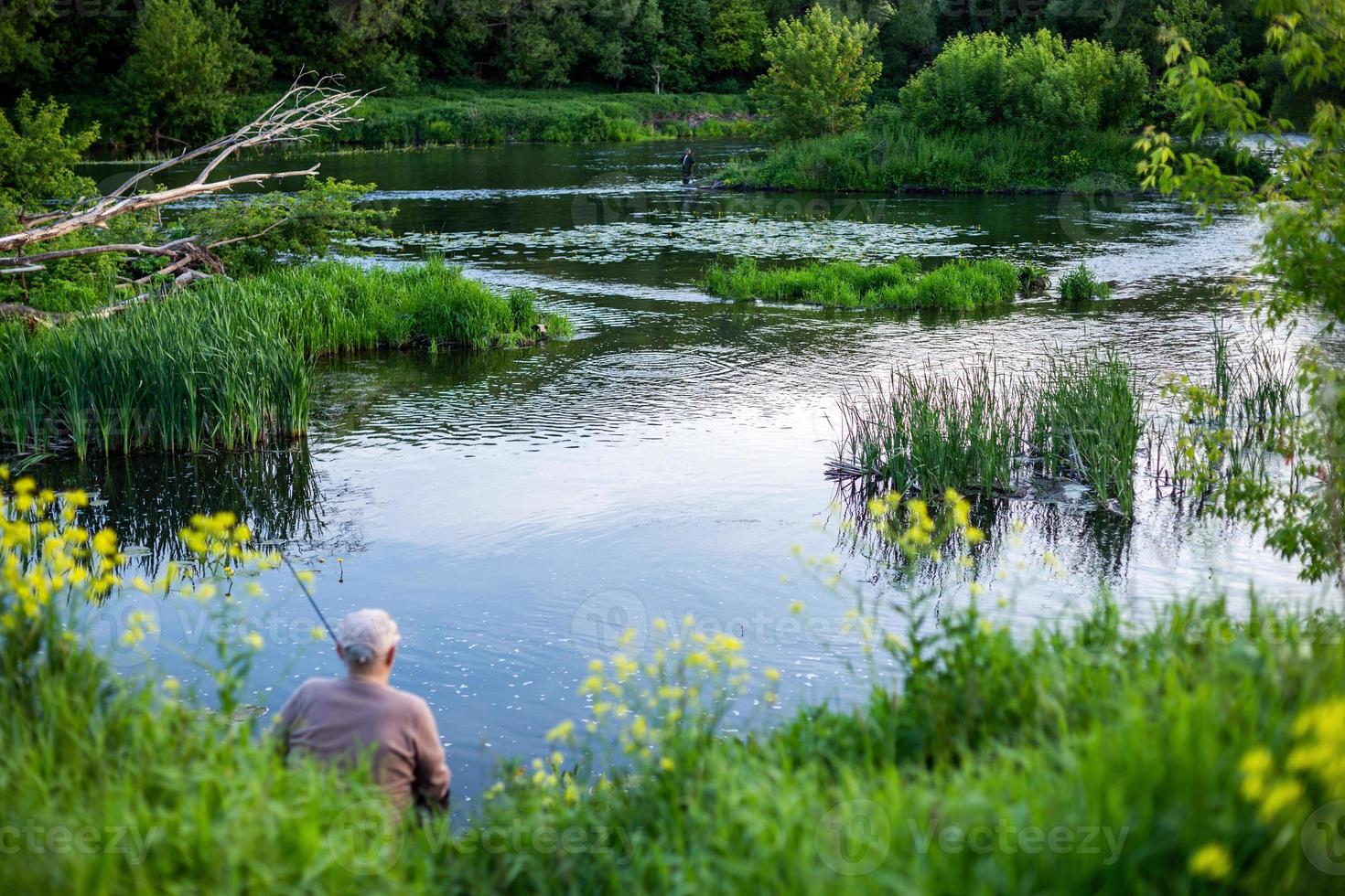 old man fishing with friend on a river at summer daytime with selective focus photo