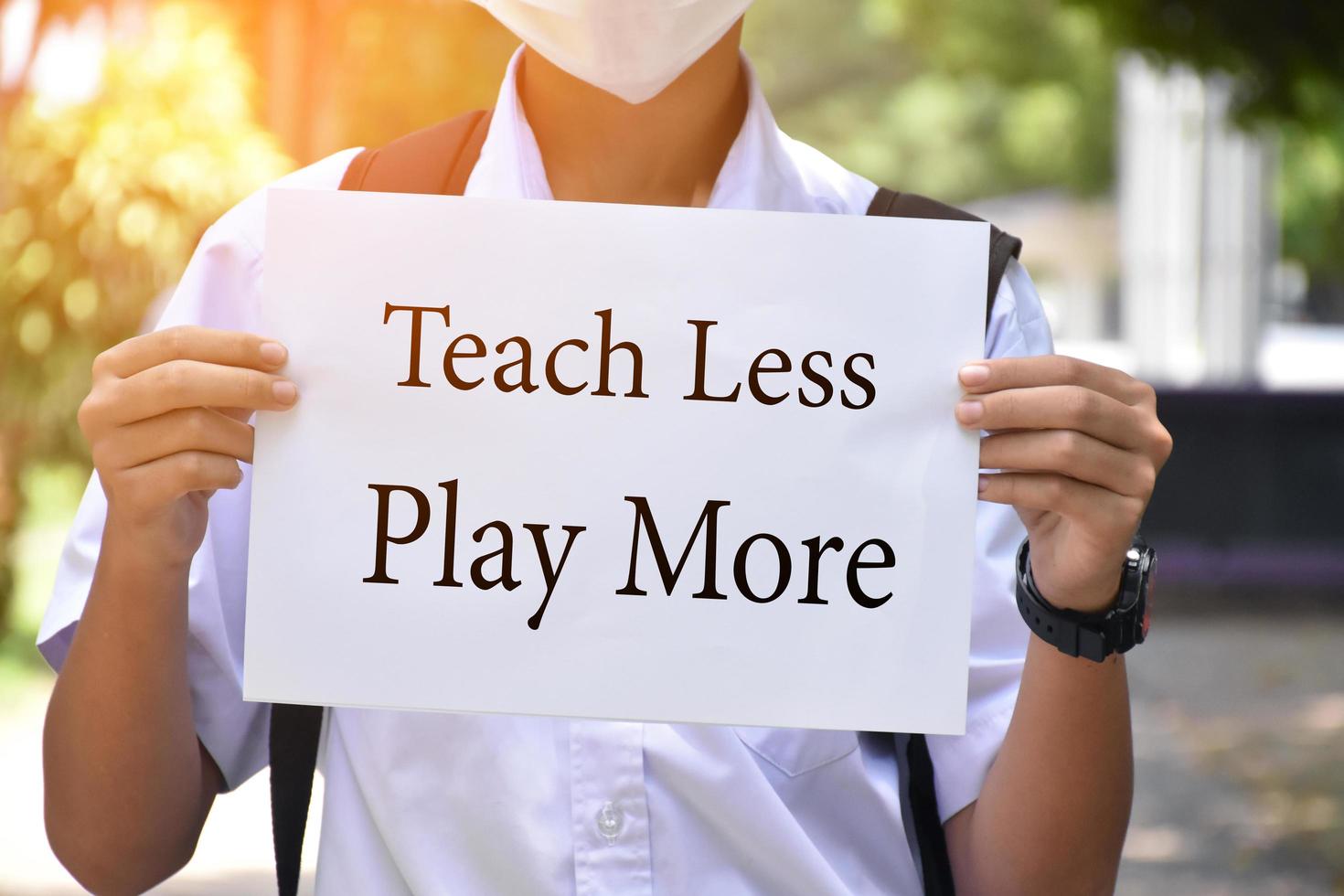 Thai boy student holds protesting paper which has text 'Teach Less Play More', soft and selective focus, concept for calling all teachers to reduce teaching in class and increase playing outclass . photo