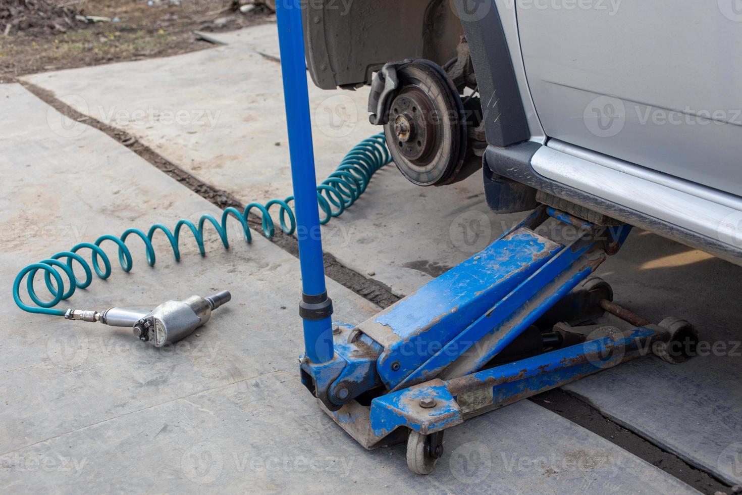 Car without wheel lifted with hydraulic floor jack and pneumatic impact wrench in a outdoor car workshop. Season tire changing procedure. photo