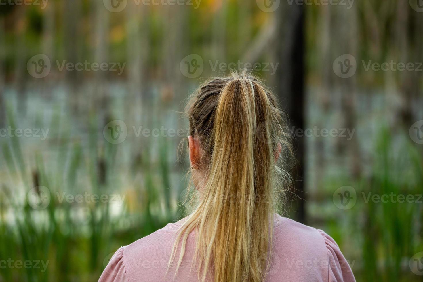 young caucasian woman standing back to camera in evening swamp with blurred dry trees and cattai in the background photo
