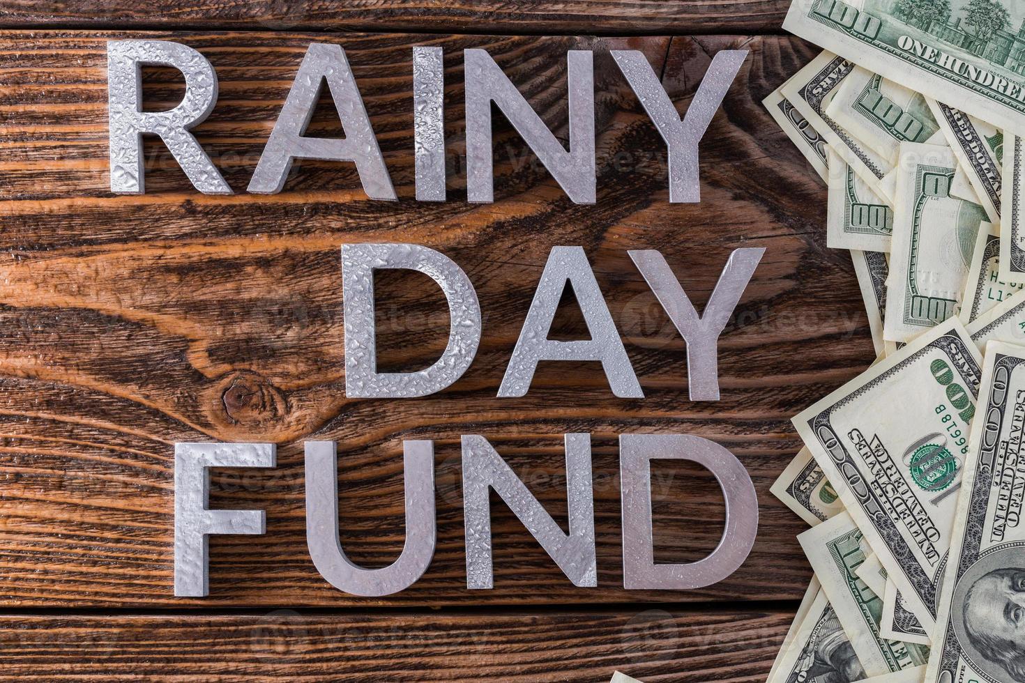 words RAINY DAY FUND laid on wooden surface by metal letters with rain drops and us dollar banknotes photo